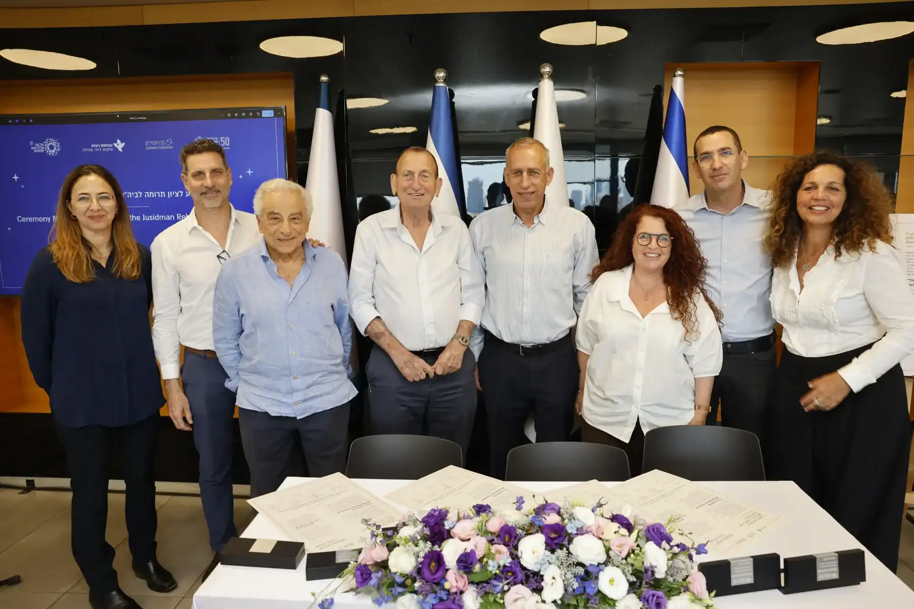 שמונה נציגים עומדים בחדר ישיבות לצד מסמכים וזר פרחים