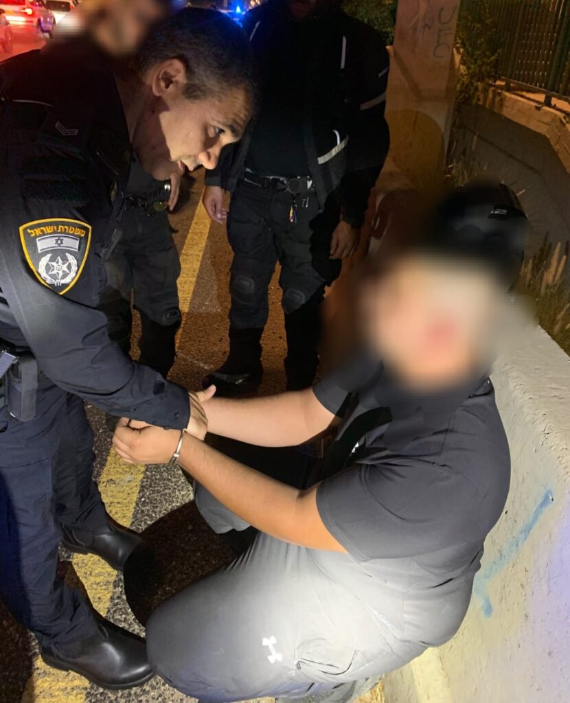 Police officers beside a suspect whose face is blurred during an arrest near a road